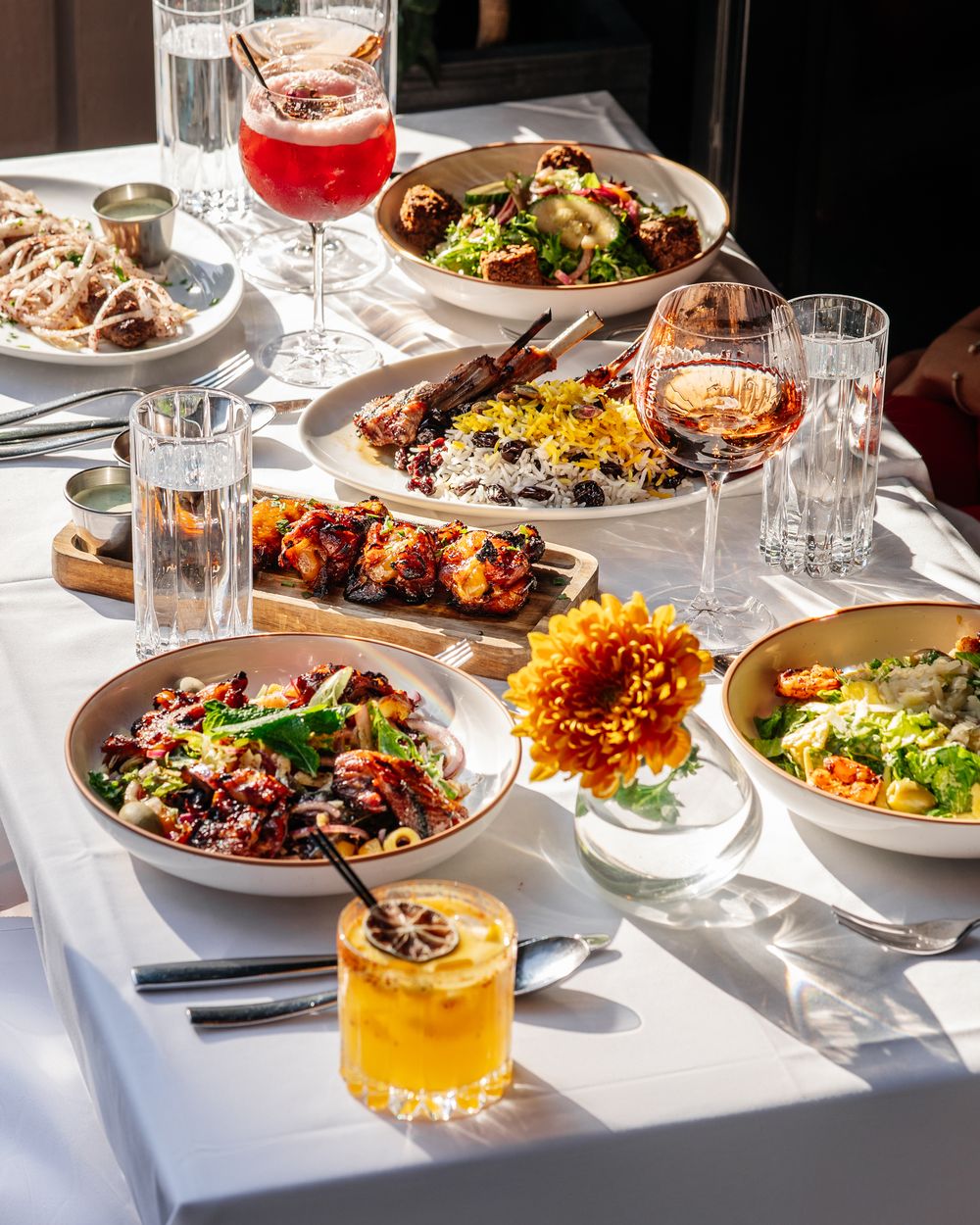 A sidewalk table of shared plates, drinks and an orange flower in Chelsea