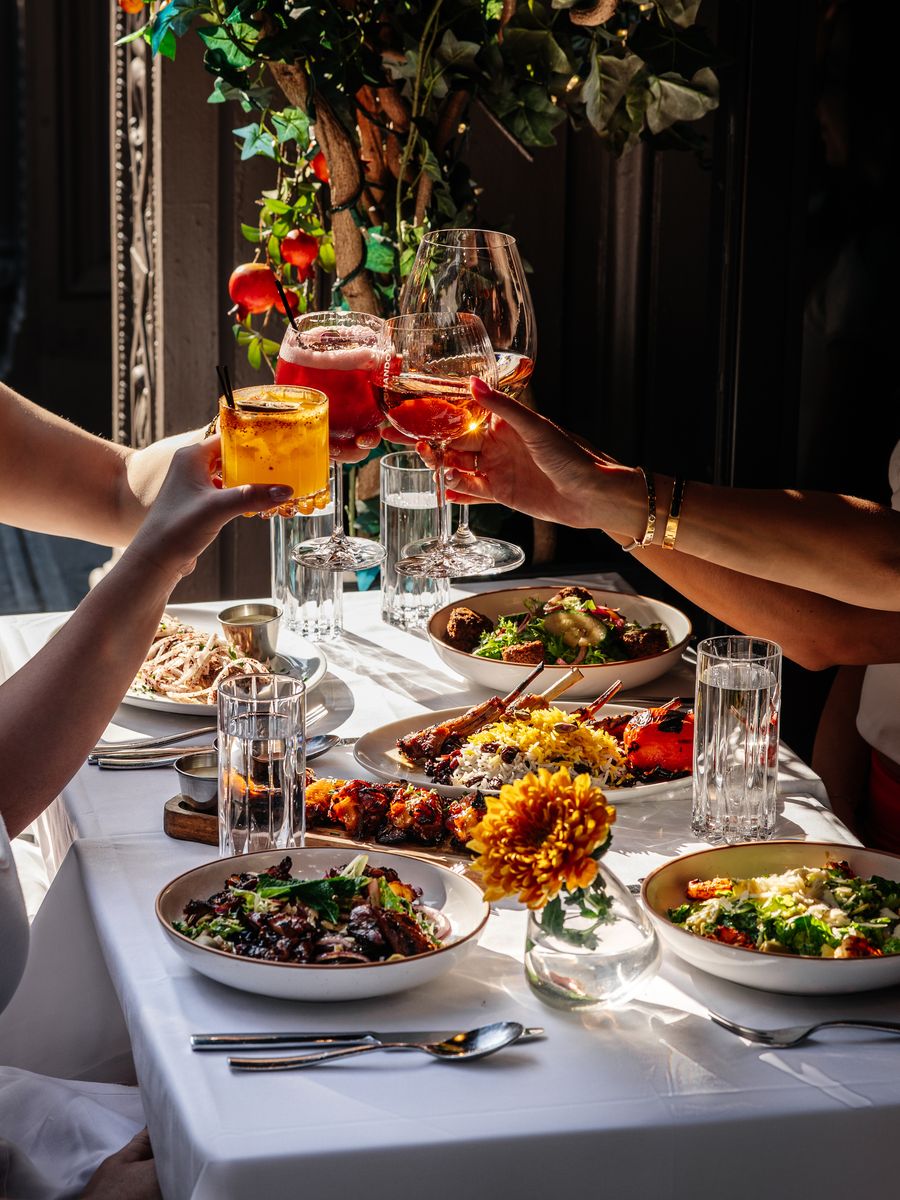 Hands clinking cocktail and wine glasses over a sidewalk table of shared plates and an orange flower in Chelsea