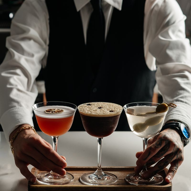 A bartender presenting three cocktails on a wooden tray